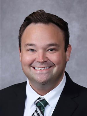 Portrait image. The individual has short, brown hair and is wearing a dark suit jacket and white shirt with a striped tie.