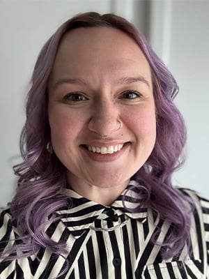 Portrait image. The individual is wearing a black and white vertically striped shirt; their hair is light, curly and shoulder length. They are smiling and looking at the camera.
