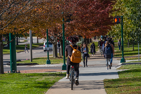 Student riding bike down the sidewalk with fall trees in the background