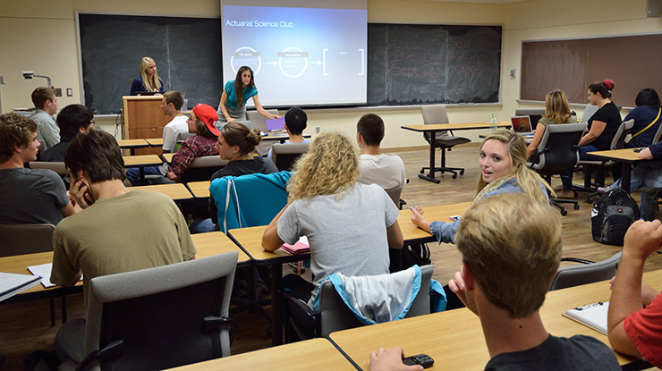 Two students make a presentation at the front of a classroom full of lively students pursuing an actuarial science degree.