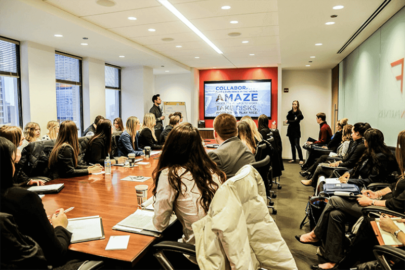 Advertising management students in formal business attire listen attentively to a presentation at the front of the room.