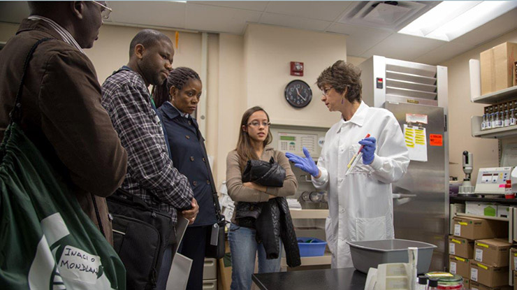 A professor in a lab coat and gloves shares information with a group of people interested in biomedical laboratory science.