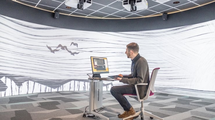 A computational data science student sits at a lone computer station surrounded by a 360? visualization space.