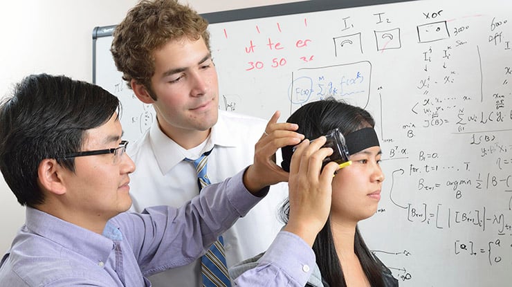 A professor affixes a computer science device around the head of a student pursuing a computer science degree.