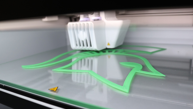 Close-up of a 3D printer in operation, printing a green Spartan helmet logo — showcasing hands-on use of engineering technology at MSU.
