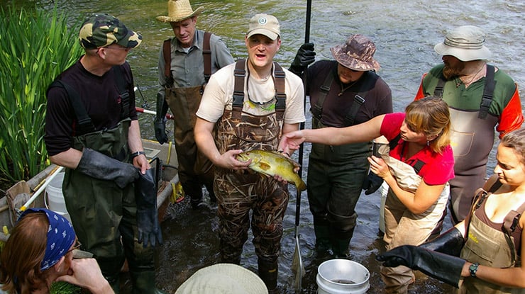 Standing in water on a sunny day, environmental biology zoology students learn from a professor holding a wild fish.