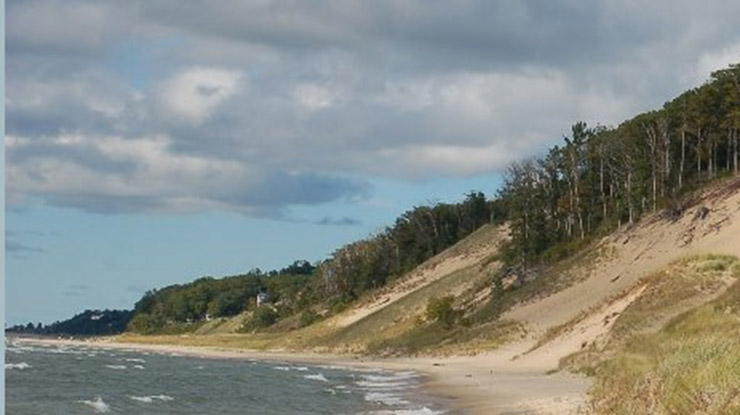 A beautiful example of environmental geography ? a hill of sand and trees overlooks a lake beneath a cloudy blue sky.