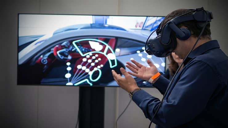 A game design degree student at Michigan State University tries out a VR headset in a room with a tv screen showing the video game.