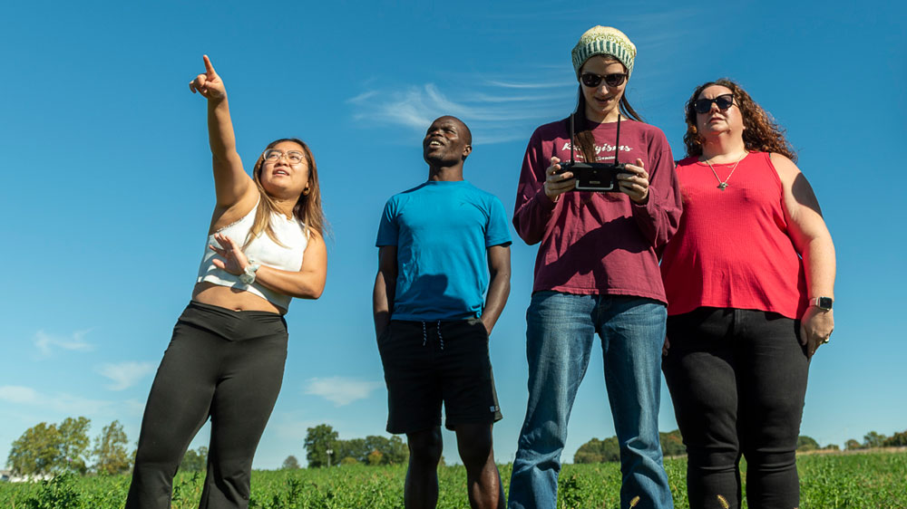 Four geography students stand together in a green field under a bright blue sky, looking up as one points toward something in the distance while another holds a drone controller.