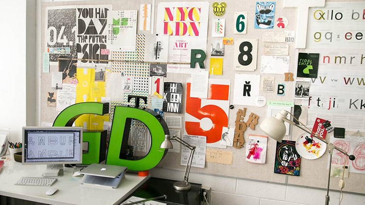 A graphic design major?s workspace ? a desk, computer and a variety of stylized letters and numbers on a bulletin board.