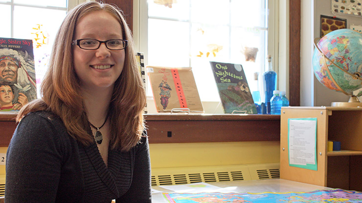 A human development and family studies major smiles and sits at a table surrounded by children?s books and a globe.