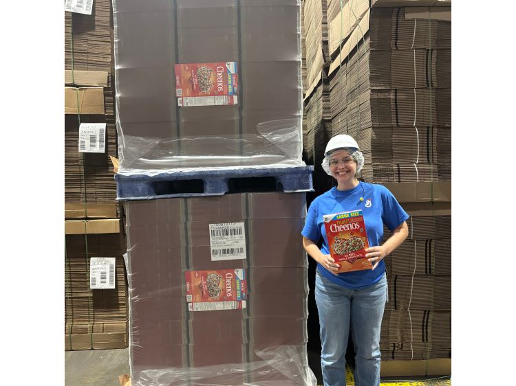 MSU student Katherine DeLorenzo smiles while holding a box of Cheerios in a warehouse, standing beside stacked pallets of cereal boxes.