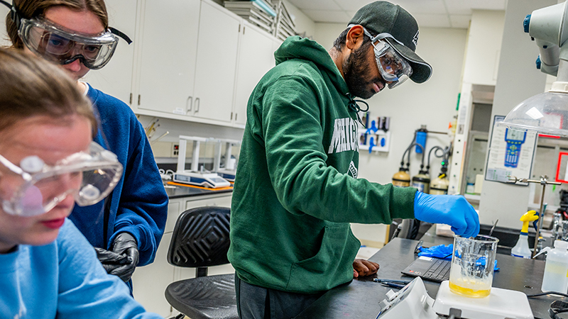 Students in safety goggles conduct a lab experiment, measuring liquid in a beaker for a physical science degree course at MSU.