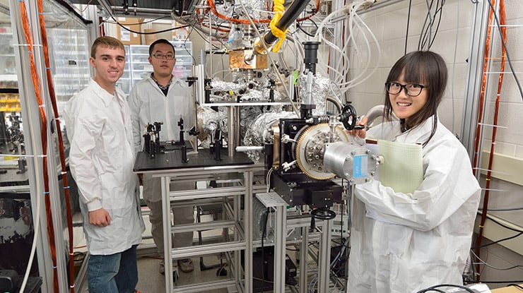 Three physics majors smile as they work in a lab with equipment that supports their learning.