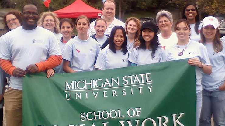A group of smiling MSU sociology majors and faculty members hold up a banner that says, ?School of Social Work.?