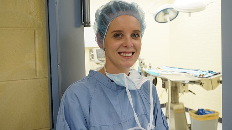 A student working on a veterinary degree wears a mask, cap and surgical gown next to an operating room.
