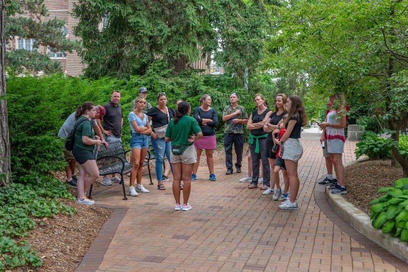 Campus tour guide speaks to a group on a brick path surrounded by trees and greenery