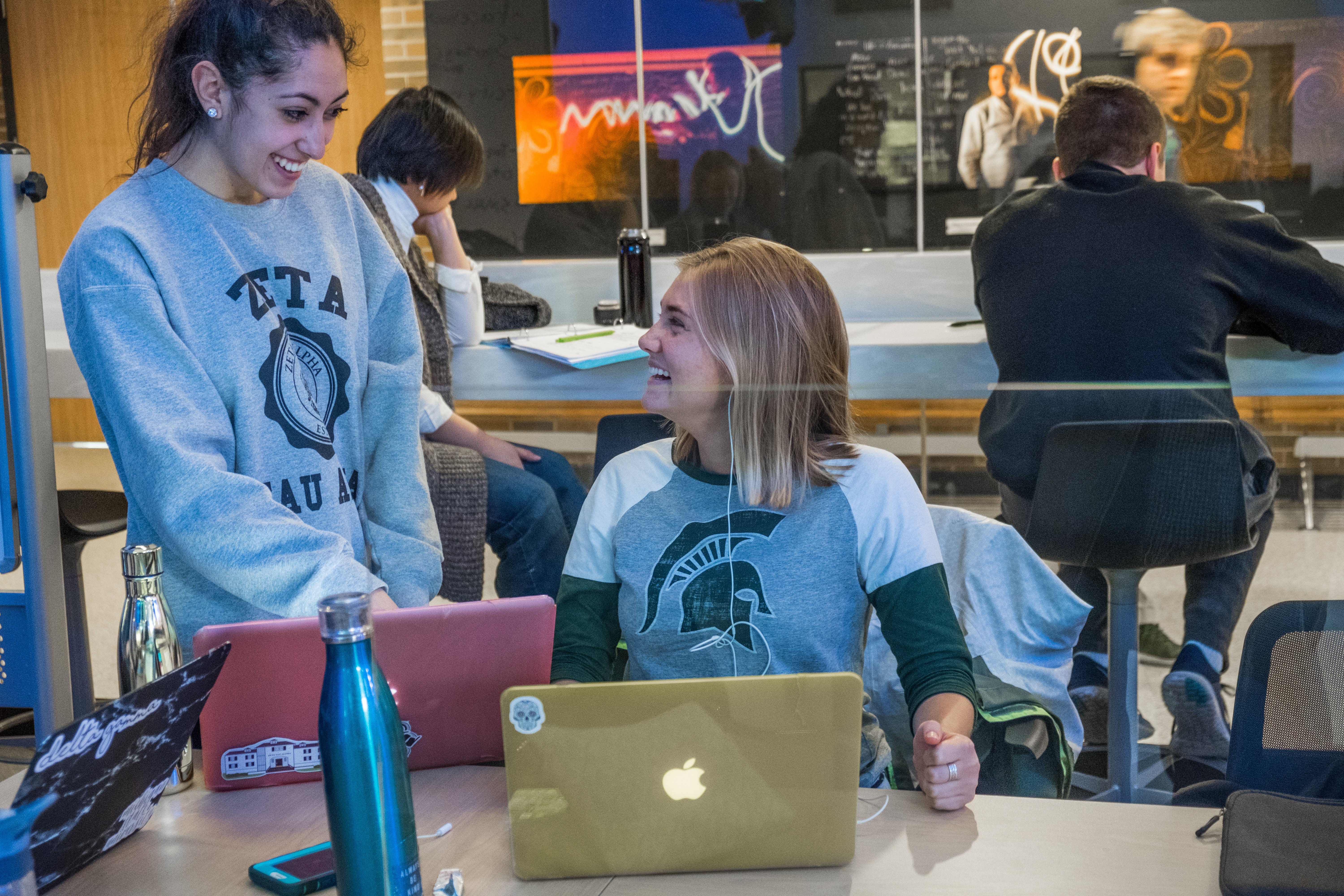 Two students with laptops smile and engage with each other in a dynamic, colorful learning space.