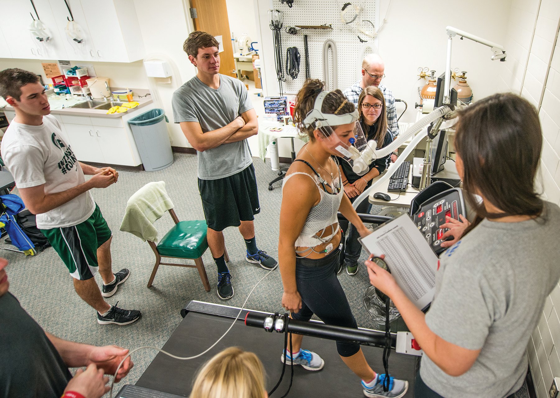 A group of students participate in a research lab while a person wearing various electrodes walks on a treadmill.