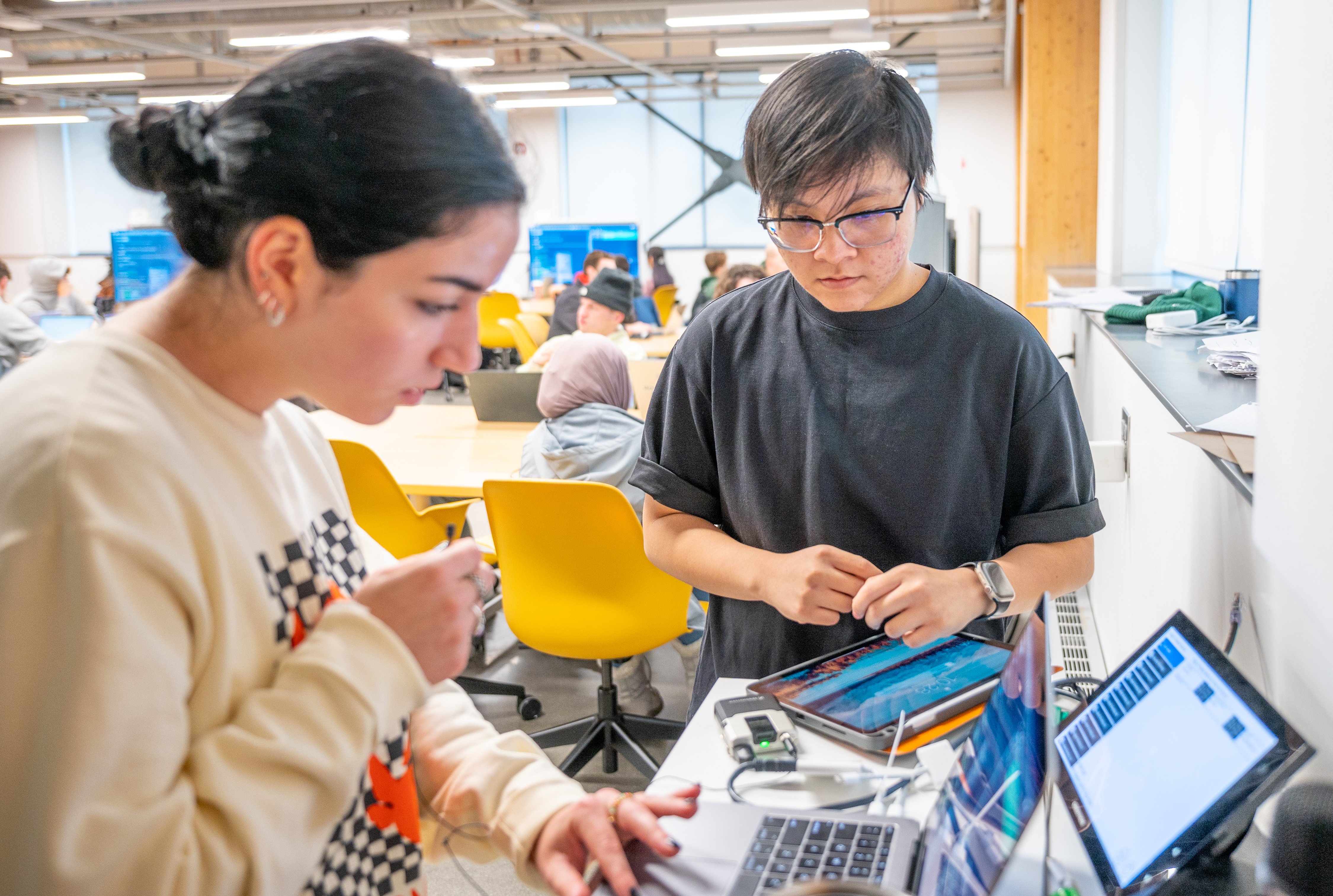 Two students work with laptops in a bright, energetic collaborative space.