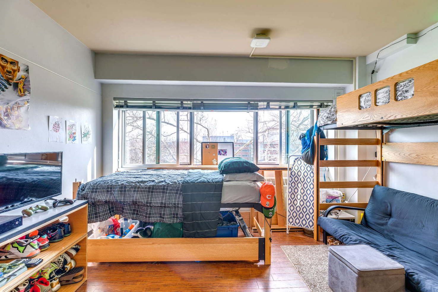 A dorm room with a bed positioned near a large window and a futon on the opposite side. A TV sits on a low stand with gaming controllers visible. The room includes storage under the bed and casual decor. Natural light fills the space, giving it a relaxed and modern feel.