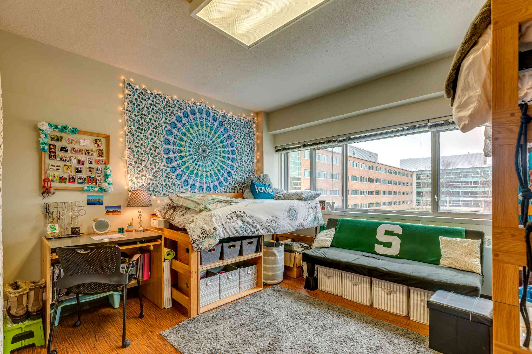 A dorm room with a raised bed next to a large window letting in natural light. The wall behind the bed is decorated with a blue patterned tapestry and string lights. A desk with personal items sits nearby, and storage bins are neatly arranged under the bed. A small couch with a green “S” blanket is positioned along the window.