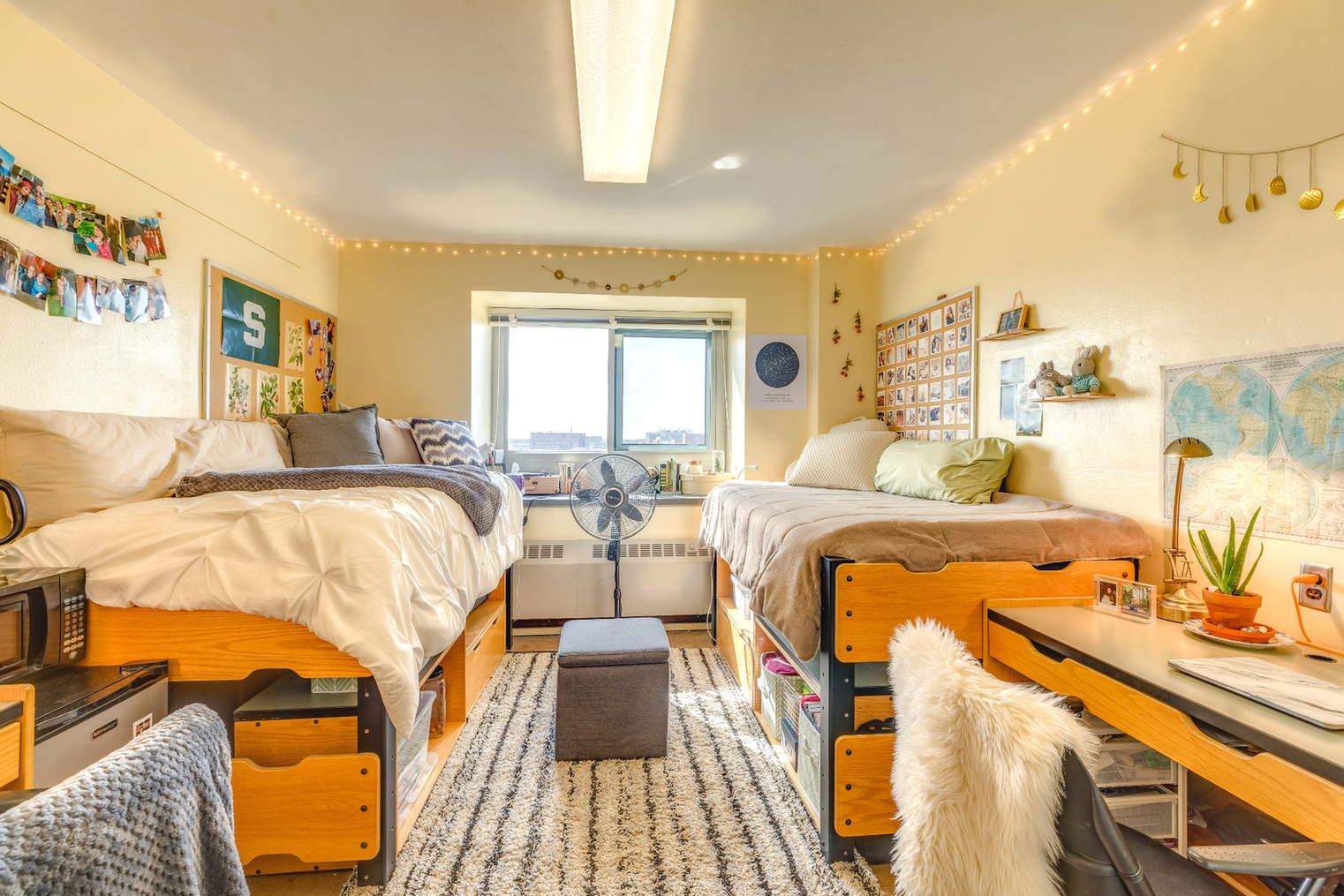 A double dorm room with two separate beds placed on opposite sides of the room. Each bed has storage drawers underneath. The room features a large window in the center, string lights along the walls, and desks on both sides. Decorations include photos, maps, and small plants, creating a bright and inviting shared space.