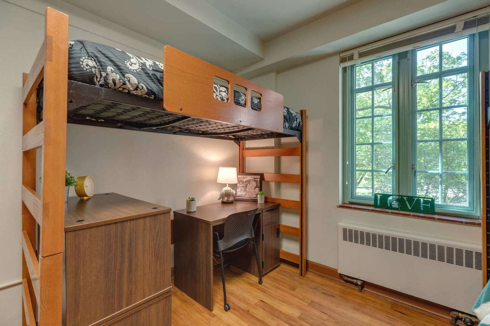 A minimally decorated dorm room with a lofted bed, a wooden desk, and a chair placed near a window. The room has neutral tones, a small lamp on the desk, and limited decorations. Natural light comes through the window, creating a clean and quiet study-focused environment.