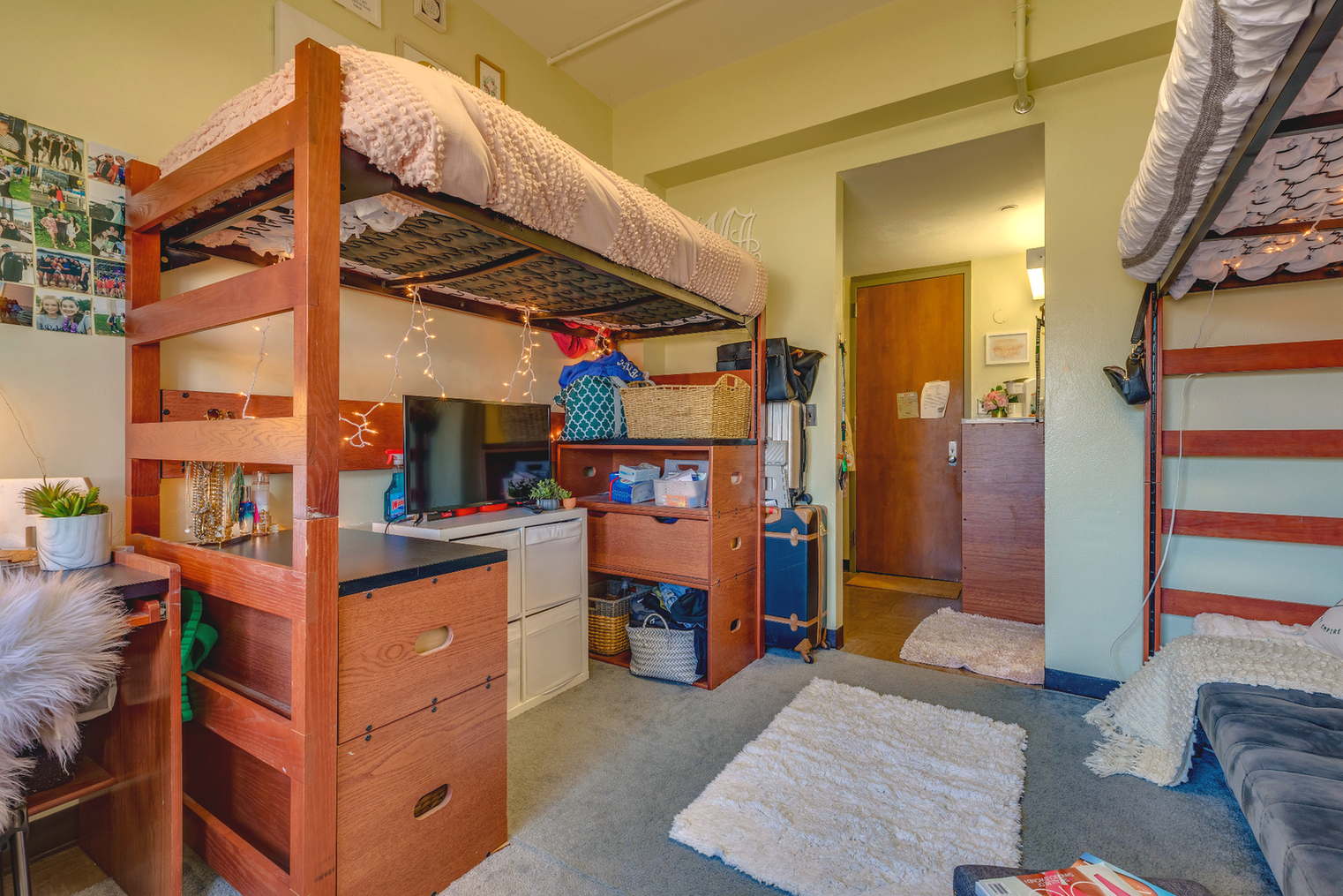 A dorm room with a lofted bed and storage drawers underneath. A small TV sits on a stand below the bed, with baskets and storage containers arranged nearby. The room includes soft rugs, string lights, and personal decor like photos on the wall. The layout emphasizes organization and efficient use of space.