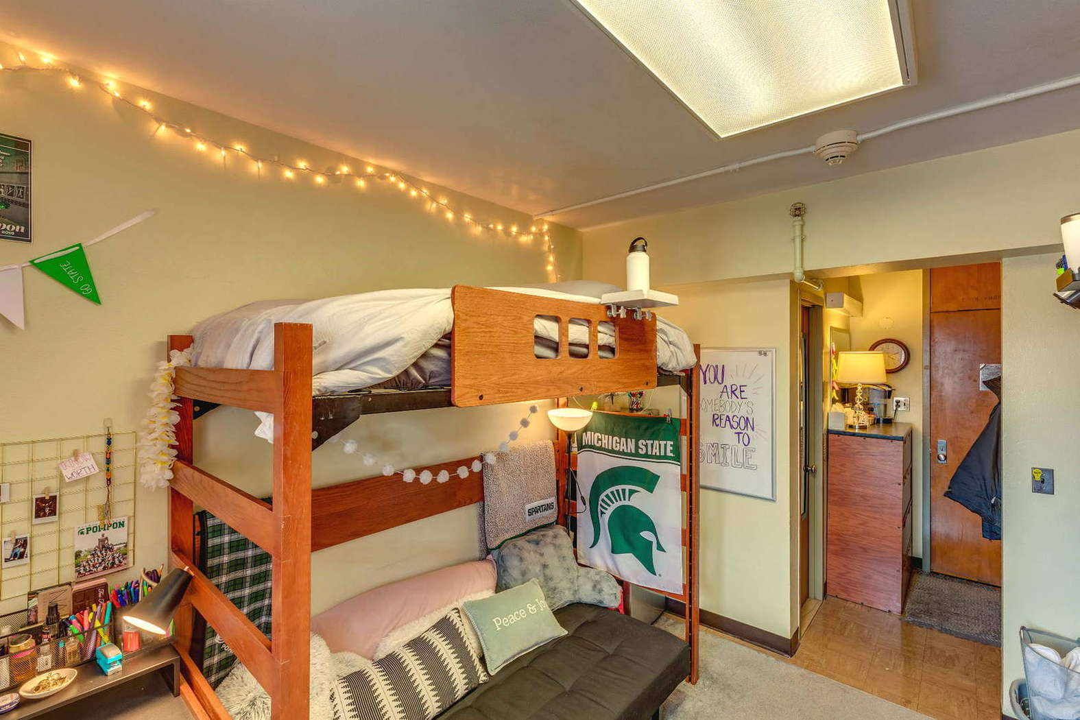 A dorm room with a lofted wooden bed and a futon underneath. The space includes string lights, a Michigan State banner, and a whiteboard with a handwritten message. A desk area with school supplies is tucked beside the bed. The room has warm lighting and a cozy, decorated feel.