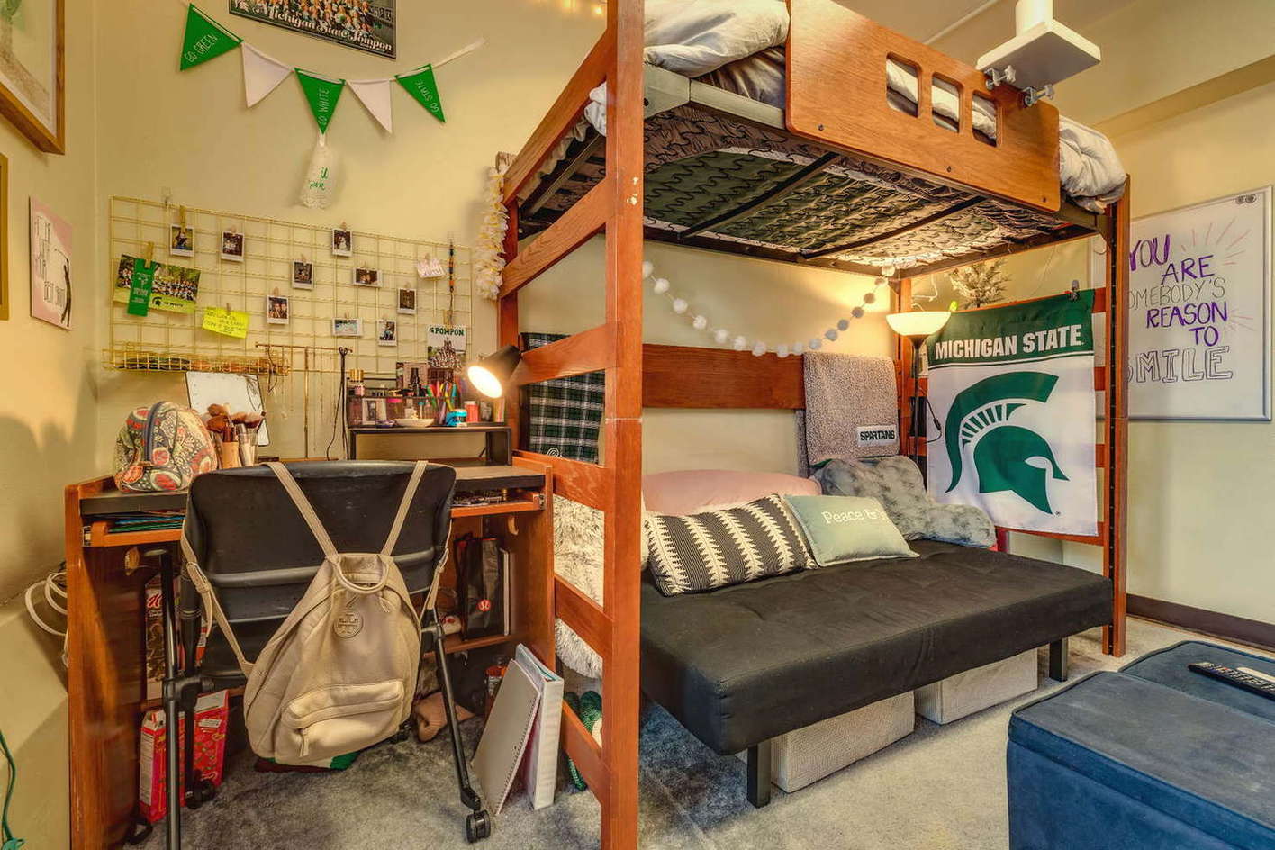 A small dorm room corner featuring a lofted wooden bed with a futon underneath. The space is decorated with Michigan State items, string lights, and a green Spartan flag. A desk with school supplies sits beside the bed, and a wall grid displays photos and keepsakes. The room feels personalized and compact.