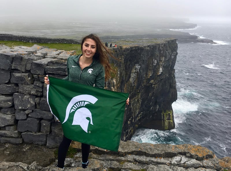 Student holds a green MSU Spartan flag on a windy, misty seaside cliff in Ireland's Aran Islands, standing by a stone wall with waves crashing below.