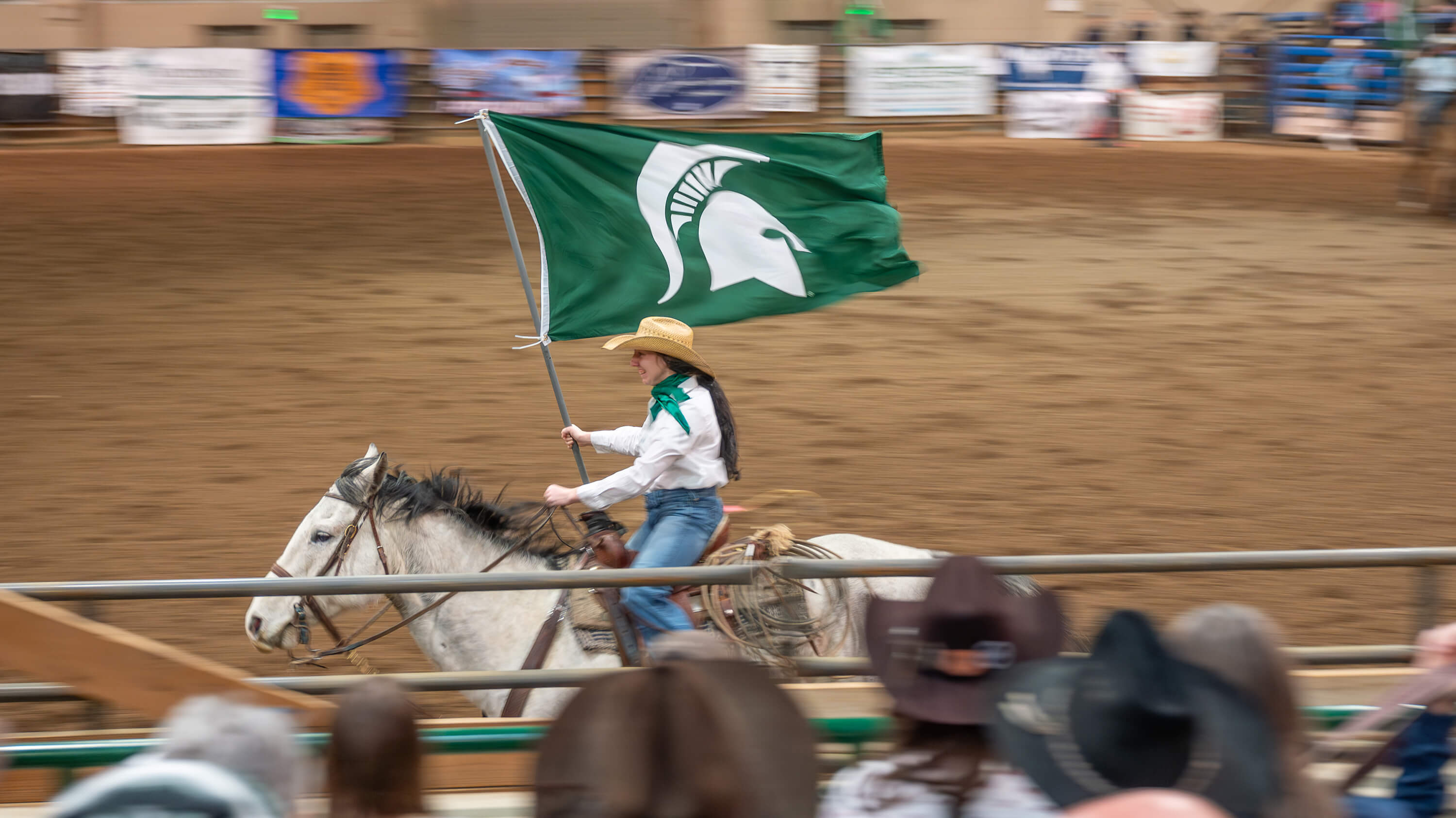 A student riding a horse and carrying a Spartan helmet flag in a rodeo event at the MSU Pavilion.