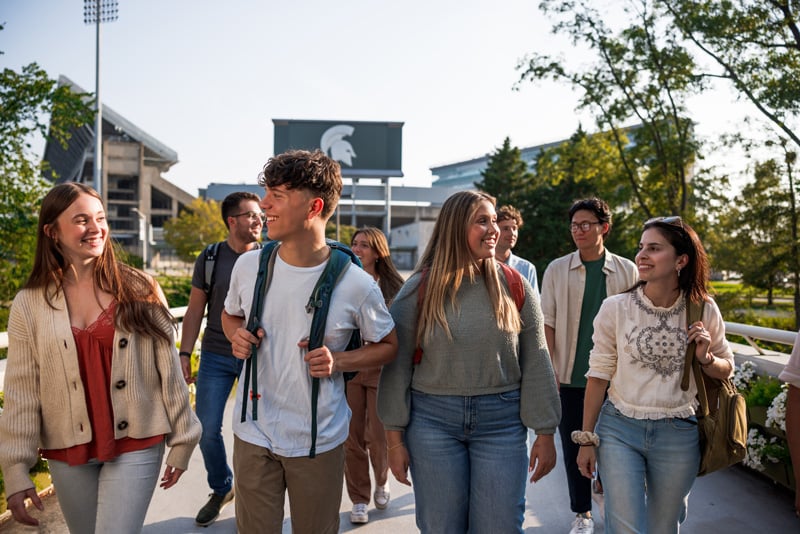 Group of Michigan State University students walk and talk on a sunny bridge over the Red Cedar River, backpacks on, with the Spartan helmet sign and stadium visible in the background.