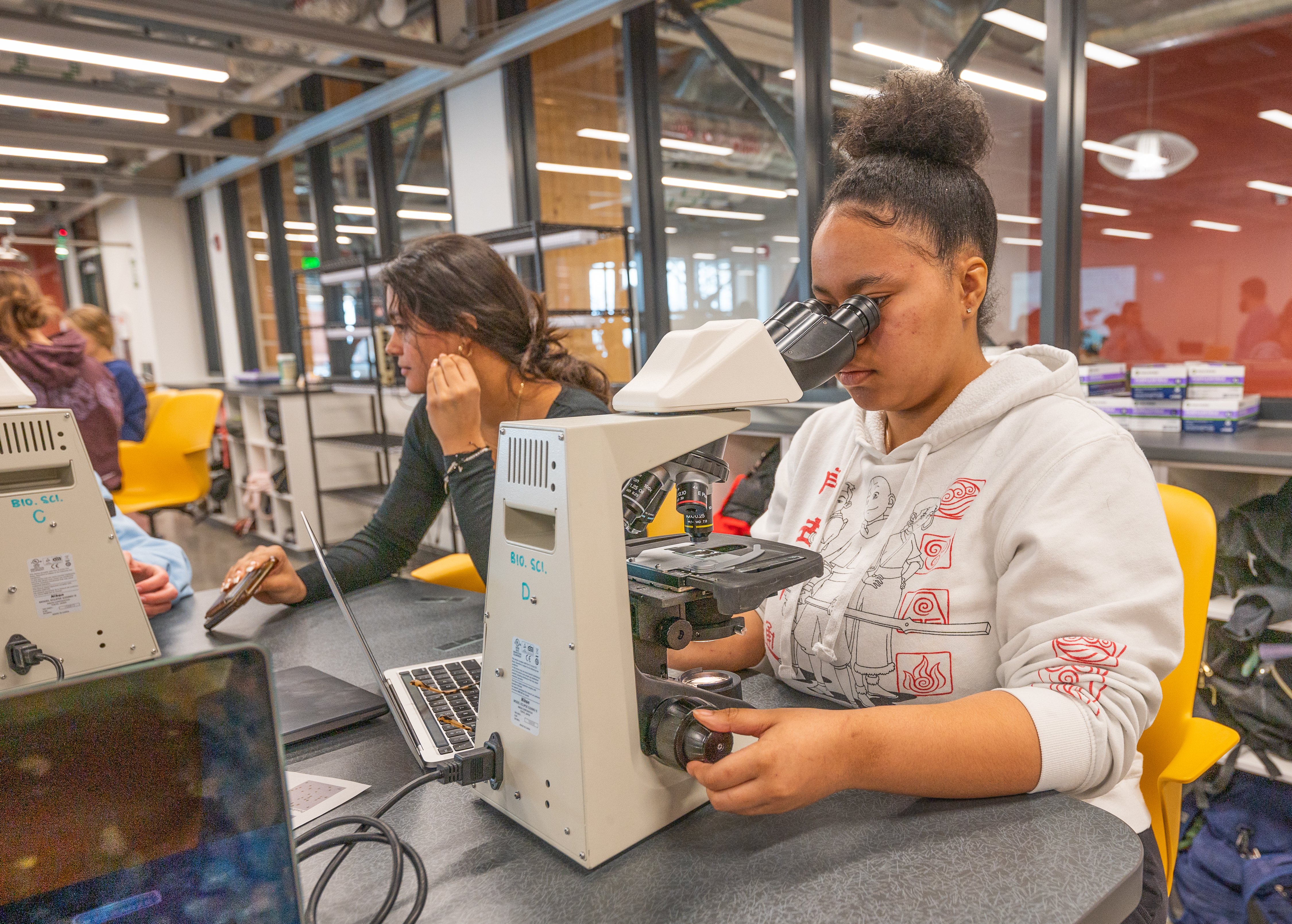 Student in a biology lab looks through a microscope while a classmate works on a laptop in a bright, modern classroom with yellow chairs and glass walls.
