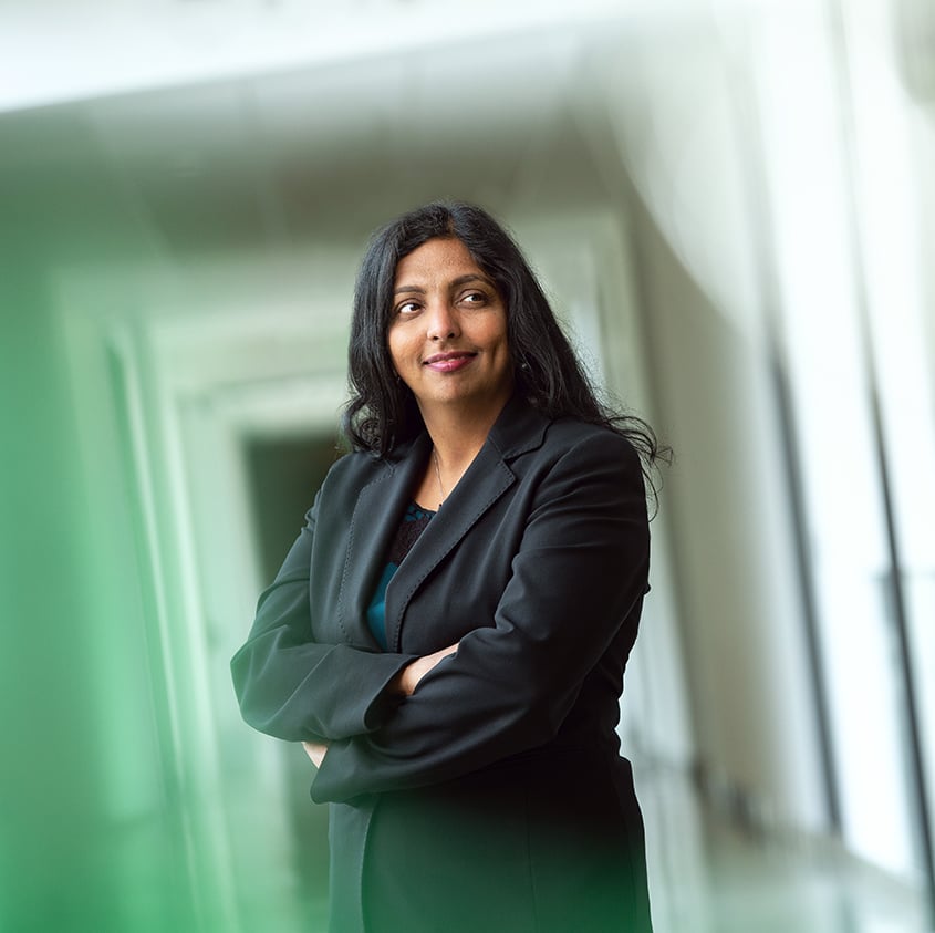 Profile picture of Anjana Susarla, smiling, standing in a brightly lit hallway with her arms crossed, wearing a dark pantsuit. 