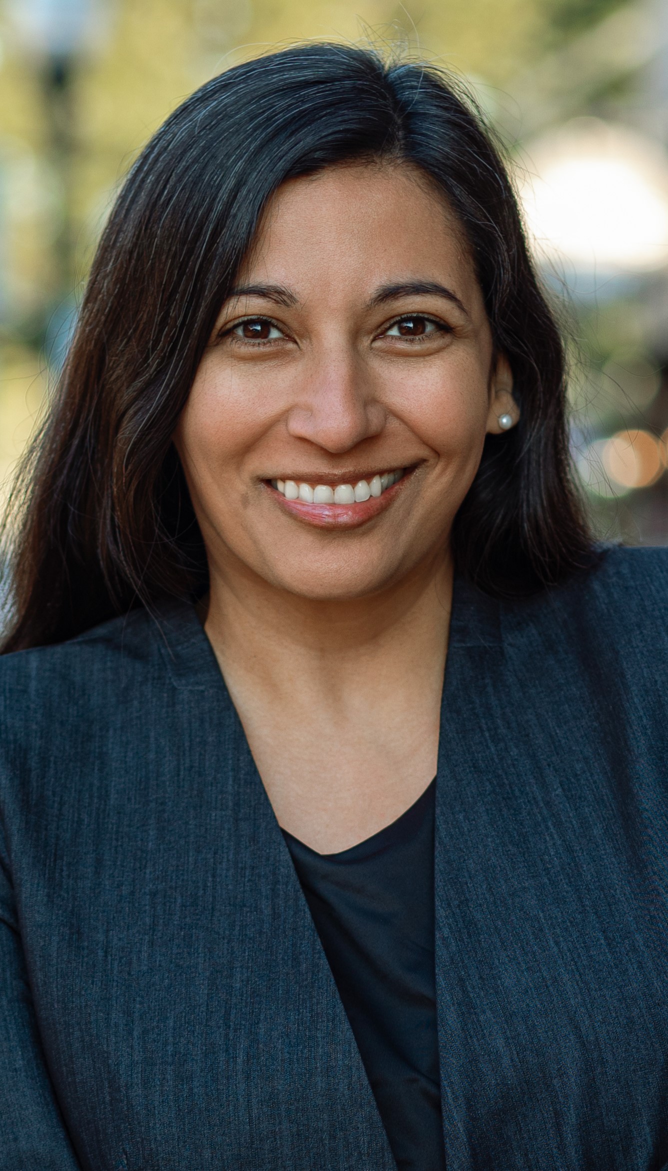 Tara Behrend, smiling, wearing a dark pant suit, standing outside with trees in the background.