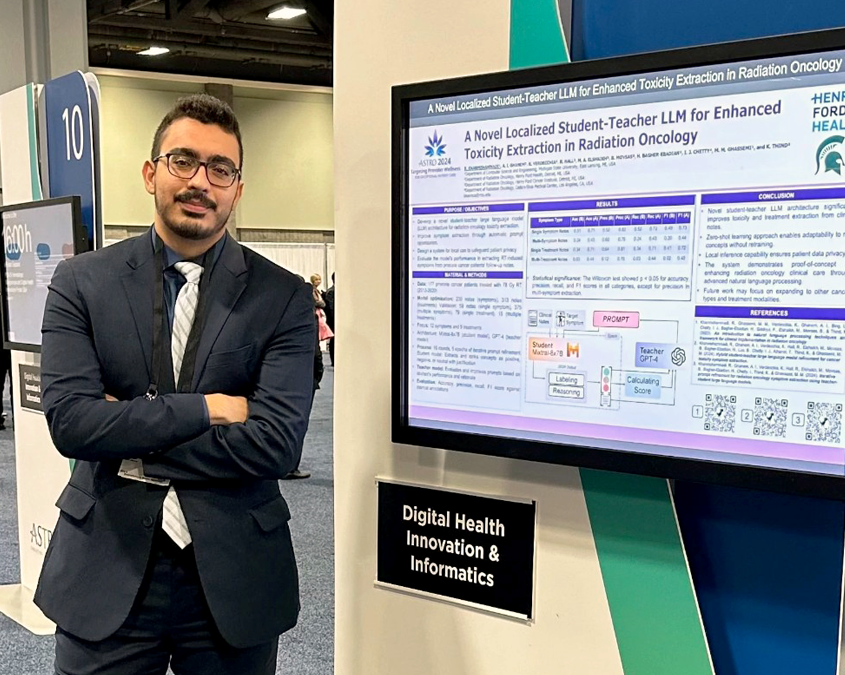 Reza Khan, wearing a dark suit and glasses, at a conference standing by a digital poster of his work.