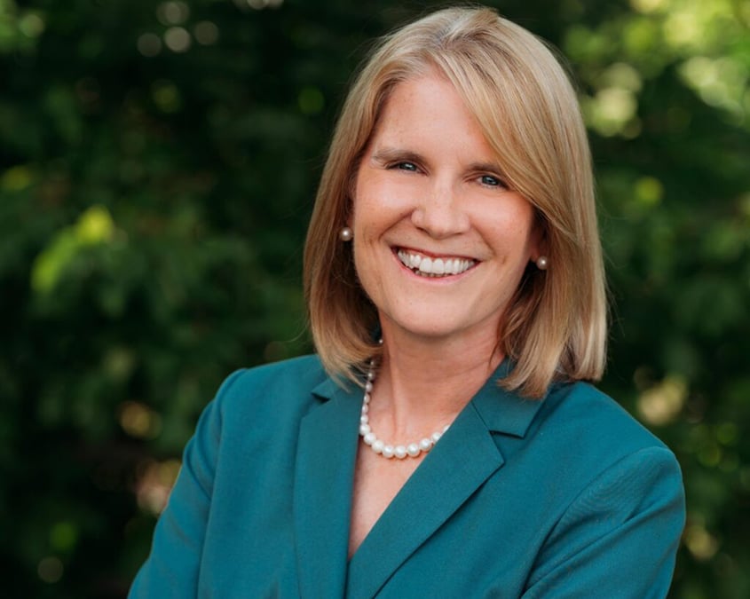 Sonja Fritzsche, smiling, wearing a turquoise blazer and pearl necklace, standing outside in front of green shrubbery.
