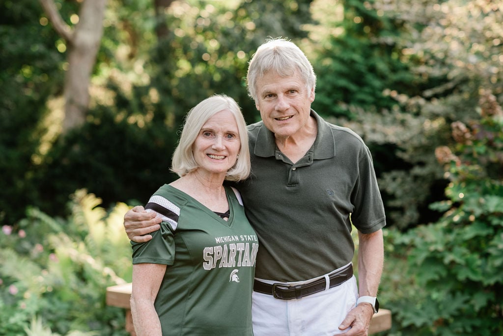 A happy couple, dressed in MSU gear, pose together in a garden setting with vibrant green foliage as the backdrop, celebrating their Spartan legacy.