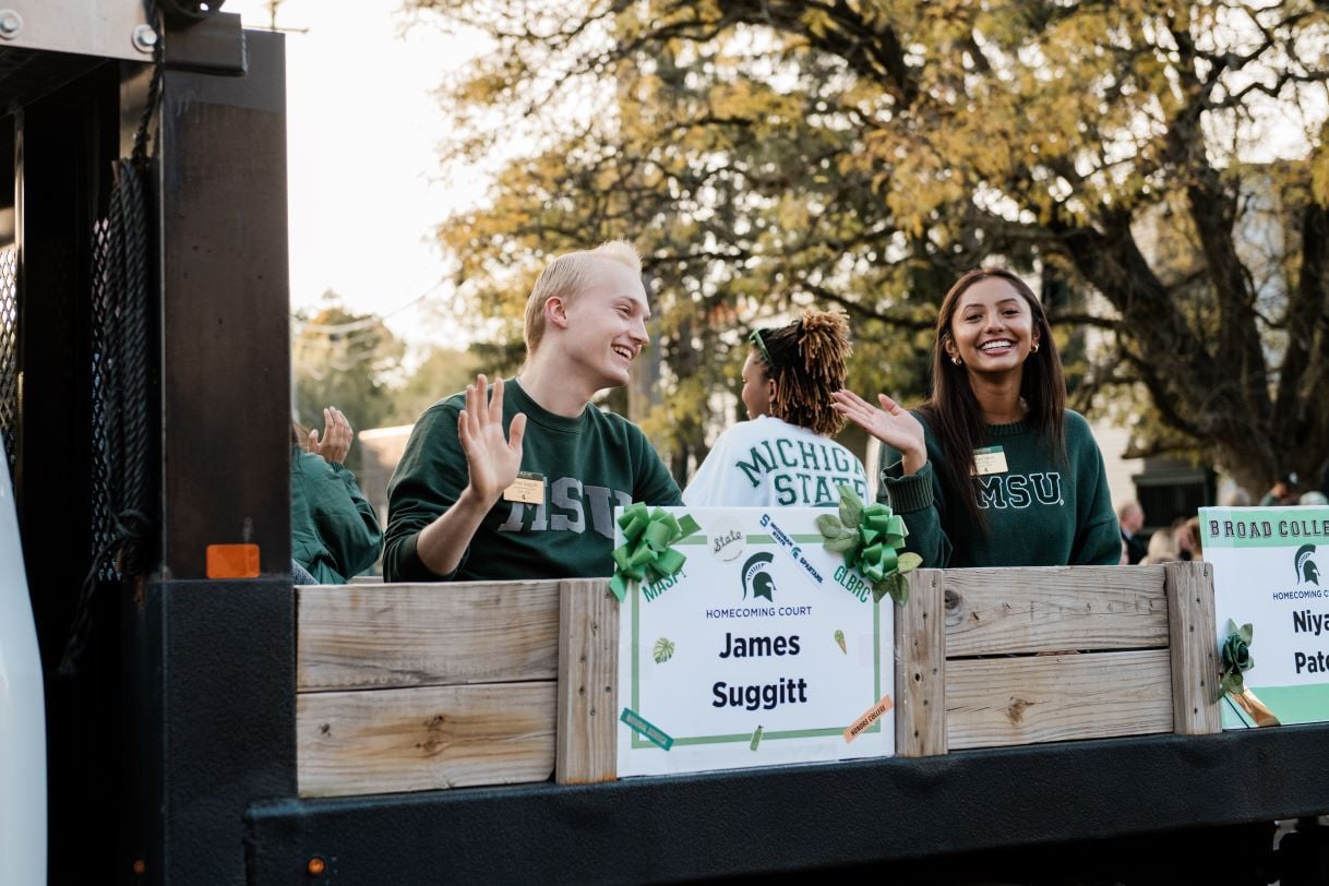 James Suggitt and a fellow member of the Michigan State University Homecoming Court wave and smile from a parade float. The float is decorated with green and white signs and surrounded by vibrant fall trees.