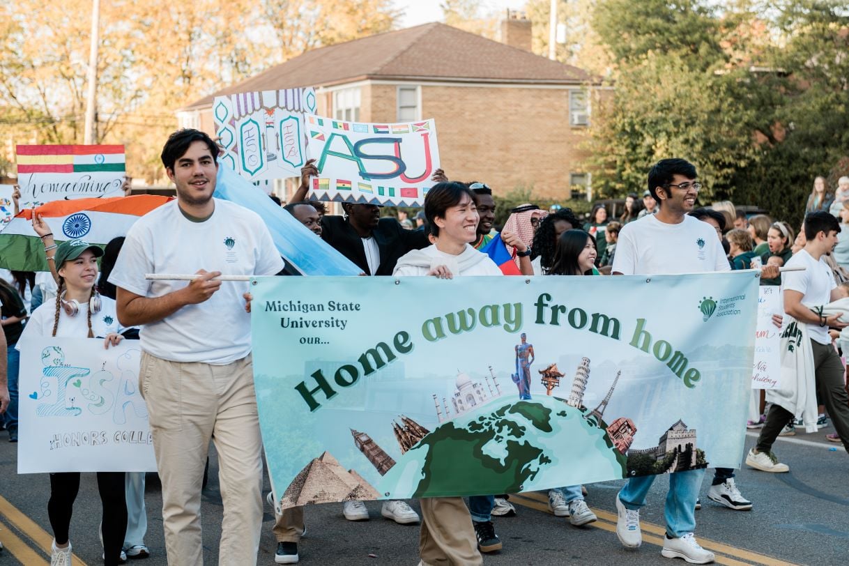 A group of Michigan State University students carry a colorful banner that reads "Home Away From Home" during the Homecoming Parade. The banner features global landmarks, celebrating international unity, with other participants holding flags and signs in the background.