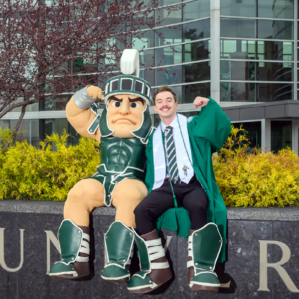 Zach O’Brien, dressed in graduation attire, poses proudly alongside the Sparty mascot in front of MSU’s campus building, both flexing their arms in celebration.