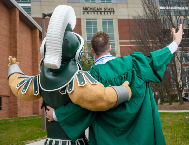 Graduate Zane Francisco and the Sparty mascot stand arm in arm, facing Spartan Stadium Tower with arms raised in celebration, capturing a proud and symbolic Spartan farewell.