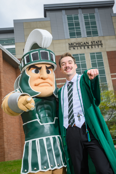 Zach O’Brien, wearing his graduation gown, stands proudly next to the Sparty mascot in front of Spartan Stadium Tower, both pointing confidently toward the camera.