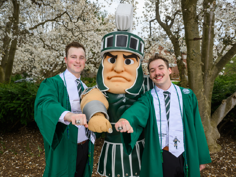Graduates Zach O’Brien and Zane Francisco pose with the Sparty mascot under blooming trees, all showing off their Sparty rings with proud fists extended toward the camera.