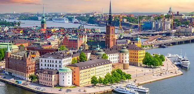 A stunning view of Stockholm, Sweden, showcasing Gamla Stan, the city's historic old town. The island is surrounded by calm waters, with colorful buildings and landmarks like Riddarholmen Church standing out. Boats are docked along the waterfront, and the cityscape extends into the distance under a vibrant sunset sky with soft orange and pink hues.