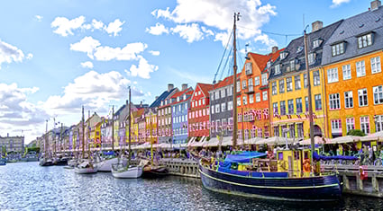 A vibrant view of Nyhavn, Copenhagen, Denmark, featuring colorful historic buildings lining a canal filled with docked boats. The water reflects the bright facades, and the Danish flags add a festive touch to the scene. People are enjoying the outdoor seating at cafés along the waterfront under a partly cloudy blue sky, creating a lively and picturesque atmosphere.