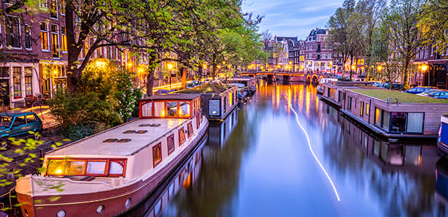 Evening view of an illuminated canal in Amsterdam, lined with houseboats, trees, and historic buildings reflecting in the water.
