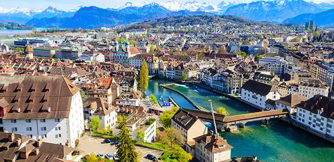 A picturesque view of Lucerne, Switzerland, featuring the historic city with its red-roofed buildings and the turquoise Reuss River running through the center. Charming wooden bridges cross the river, including the iconic Chapel Bridge. The city is surrounded by rolling hills and snow-capped mountains under a clear blue sky, creating a stunning backdrop.